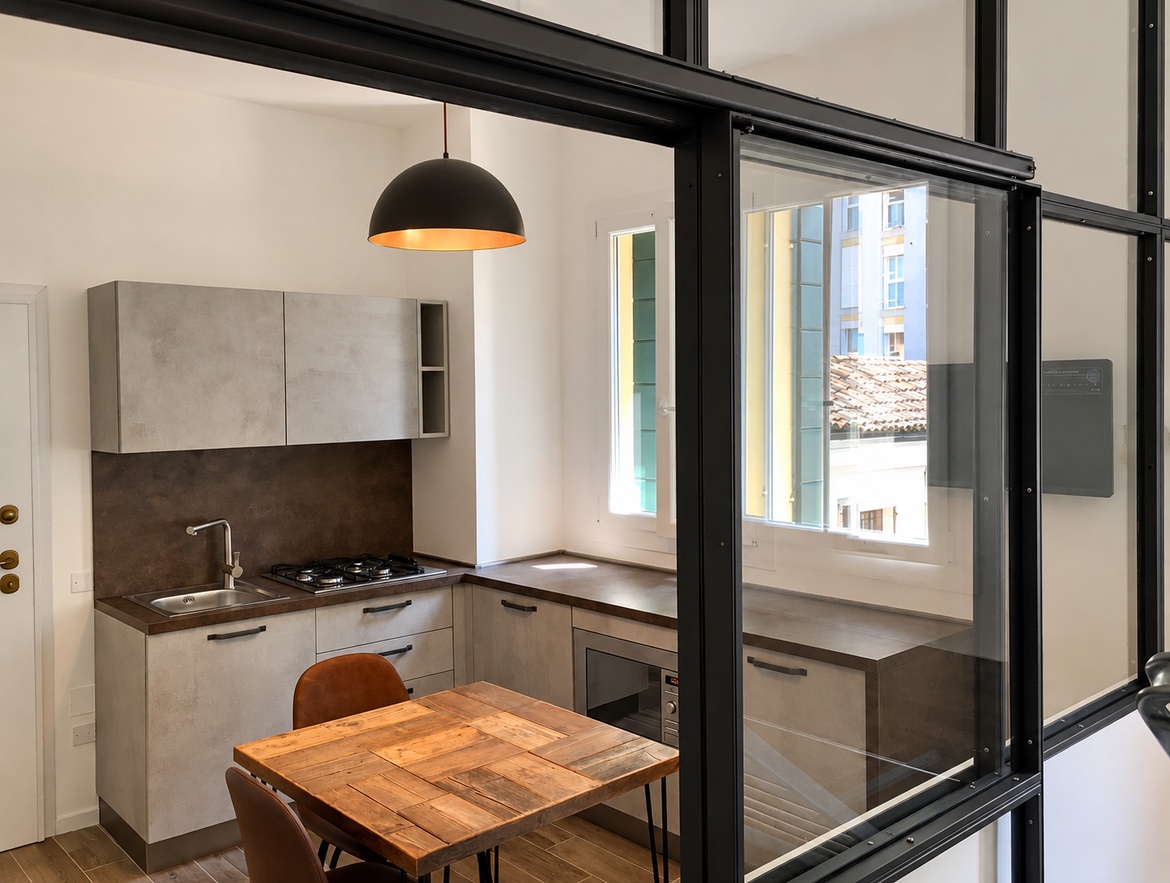 Cucina su misura con finiture effetto cemento e ruggine, tavolo in legno e vetrata in ferro e vetro nell’Appartamento Via Dante a Padova.